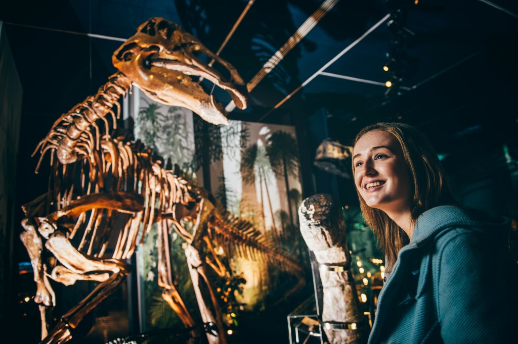 Young women posing for a picture with an impressive dinosaur display, located in Cardiff Museum.